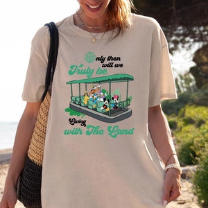 May include: A woman wearing a light beige t-shirt with a green graphic of a boat with the text "Only then will we truly be going with the land".