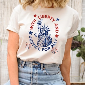 May include: A cream-colored short-sleeve t-shirt featuring a blue graphic of the Statue of Liberty centered on the chest. The design includes red and blue stars, firework motifs, and the text "LIBERTY AND JUSTICE FOR ALL" in a circular layout.