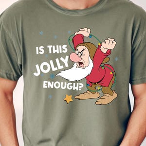 May include: Olive green t-shirt featuring a cartoon image of Grumpy from Snow White, holding Christmas lights and the text "IS THIS JOLLY ENOUGH?". The design includes snowflakes and a gold star.