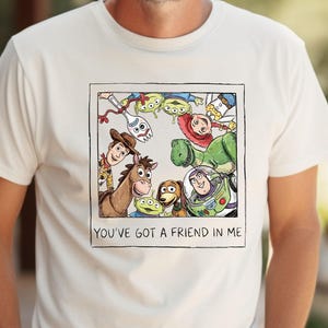 Puede incluir: Camiseta color crema con un gr&aacute;fico enmarcado de personajes de Toy Story, incluyendo a Woody, Buzz Lightyear y los extraterrestres. El texto "YOU'VE GOT A FRIEND IN ME" est&aacute; impreso debajo de la imagen. La camiseta la lleva una persona.