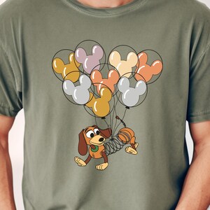 May include: A green t-shirt with a cartoon dog holding a bunch of colorful Mickey Mouse balloons. The dog is wearing a spring-like body and has a brown and white spotted pattern.