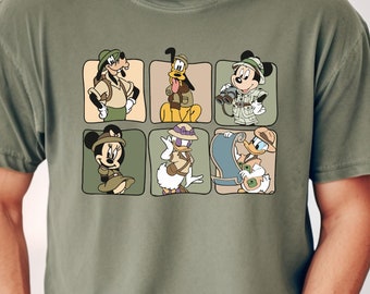 May include: A green t-shirt featuring a cartoon image of Mickey Mouse, Minnie Mouse, Donald Duck, Goofy, and Pluto dressed in safari outfits. The characters are arranged in a grid pattern.