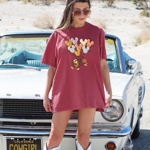 May include: A woman wearing a maroon t-shirt with a cartoon dog and balloons printed on it. She is standing in front of a white vintage car with a "California Cowgirl" license plate.