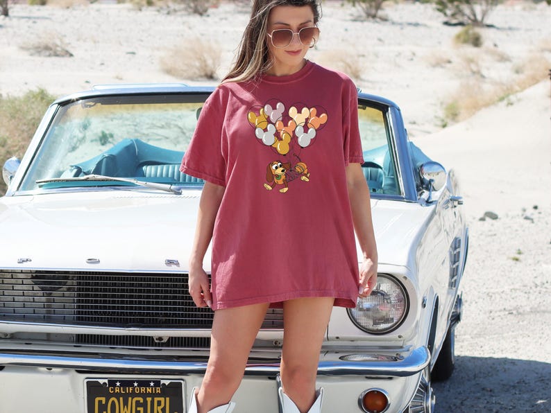 May include: A woman wearing a maroon t-shirt with a cartoon dog and balloons printed on it. She is standing in front of a white vintage car with a "California Cowgirl" license plate.
