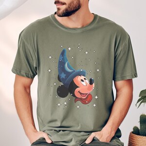 May include: Olive green t-shirt featuring a cartoon Mickey Mouse wearing a blue wizard hat with stars and a crescent moon. The design includes a red scarf and is surrounded by white star graphics.