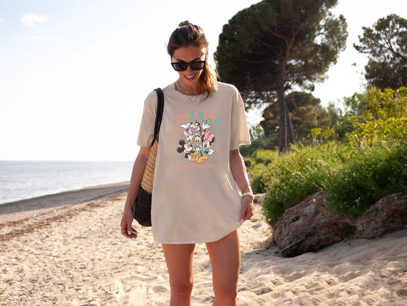 May include: A woman wearing a beige t-shirt with a colorful graphic of Mickey Mouse and friends. The graphic says "Disney Parks" and "California Adventure".