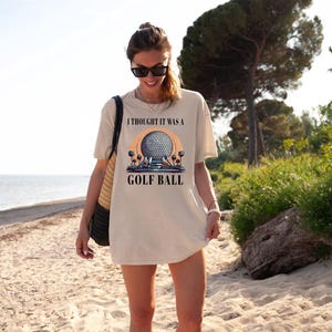 Puede incluir: Una mujer con una camiseta beige con un gr&aacute;fico de una bola de golf y el texto "I thought it was a golf ball". Est&aacute; de pie en una playa de arena.