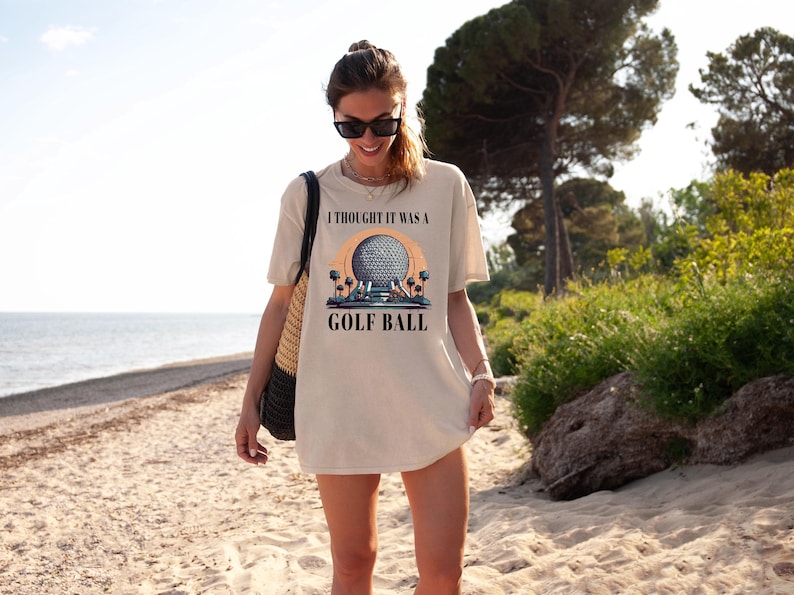 Puede incluir: Una mujer con una camiseta beige con un gr&aacute;fico de una bola de golf y el texto "I thought it was a golf ball". Est&aacute; de pie en una playa de arena.