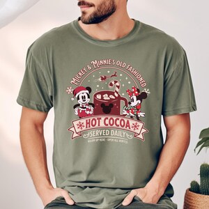 May include: Olive green t-shirt featuring a graphic of Mickey and Minnie Mouse with the text "Mickey & Minnie's Old Fashioned Hot Cocoa Served Daily". The design includes a mug of cocoa, snowflakes, and a candy cane.