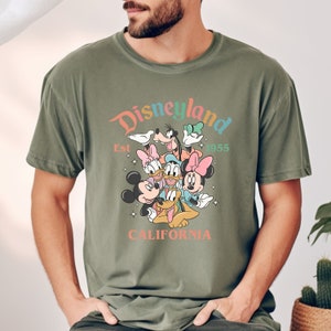 May include: A green t-shirt with a vintage-style graphic of Mickey Mouse and friends at Disneyland. The text reads "Disneyland Est 1955 California".