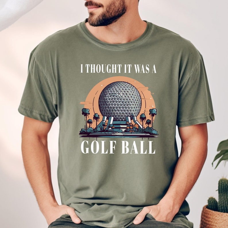 Puede incluir: Una camiseta verde con el texto "I thought it was a golf ball" y una imagen de dibujos animados de una gran esfera con un patr&oacute;n geom&eacute;trico, rodeada de palmeras.