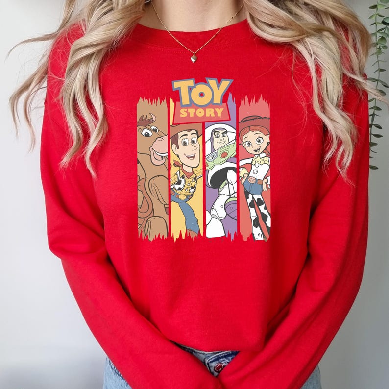 May include: A red sweatshirt featuring the Toy Story logo and character illustrations. The design includes images of Bullseye, Woody, Buzz Lightyear, and Jessie. The word "TOY" is in blue and yellow, and "STORY" is in orange.