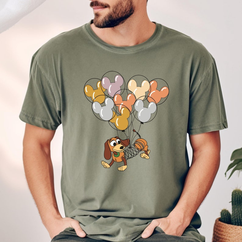 May include: A green t-shirt with a cartoon dog holding a bunch of colorful Mickey Mouse balloons. The dog is wearing a spring-like body and has a brown and white spotted pattern.
