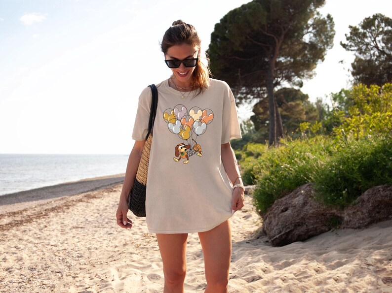 May include: A woman wearing a beige t-shirt with a cartoon dog holding balloons in the shape of hearts. The dog is brown and white and the balloons are various shades of pink, orange, and yellow.