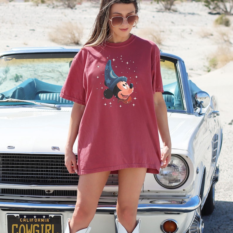 May include: A maroon t-shirt featuring a cartoon Mickey Mouse in a wizard hat with stars. The graphic tee is worn by a person standing near a classic white car. The shirt is a casual, oversized fit.