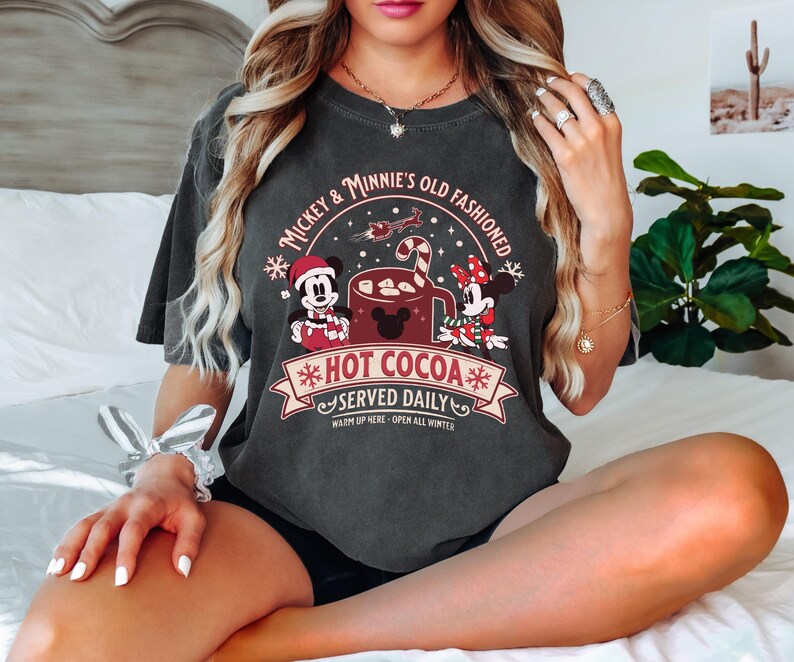 May include: Dark gray t-shirt featuring a graphic of Mickey and Minnie Mouse with a hot cocoa design. The text reads "Mickey & Minnie's Old Fashioned Hot Cocoa Served Daily. Warm up here - Open all winter."