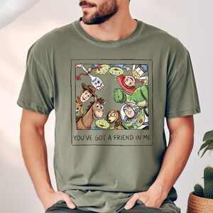 Puede incluir: Camiseta verde oliva con un gr&aacute;fico enmarcado de personajes de Toy Story. El gr&aacute;fico incluye a Woody, Buzz Lightyear, Rex y otros. El texto "YOU'VE GOT A FRIEND IN ME" est&aacute; debajo.