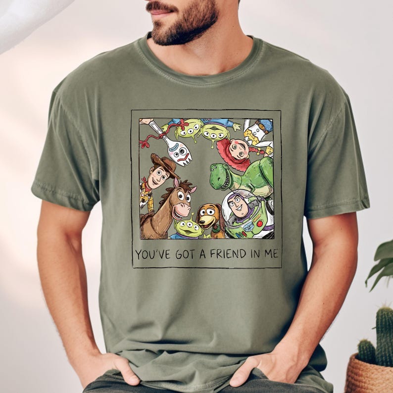 Puede incluir: Camiseta verde oliva con un gr&aacute;fico enmarcado de personajes de Toy Story. El gr&aacute;fico incluye a Woody, Buzz Lightyear, Rex y otros. El texto "YOU'VE GOT A FRIEND IN ME" est&aacute; debajo.