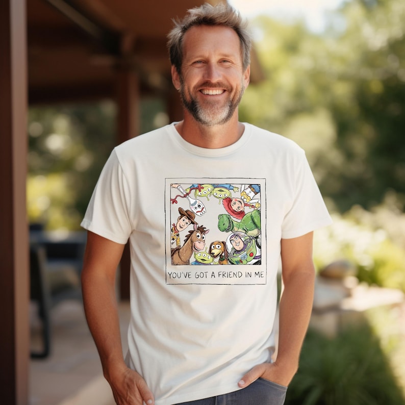 Puede incluir: Camiseta color crema con un gr&aacute;fico enmarcado de personajes de Toy Story, incluyendo a Woody, Buzz Lightyear y los extraterrestres. El texto "YOU'VE GOT A FRIEND IN ME" est&aacute; impreso debajo de la imagen. La camiseta la lleva una persona.