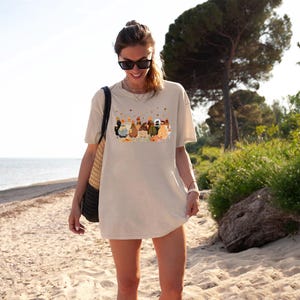May include: A woman wearing a beige t-shirt with a graphic print of a group of chickens and the text "Cluckin' Crazy". She is standing on a sandy beach with a straw bag over her shoulder.