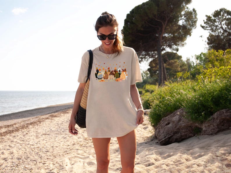 May include: A woman wearing a beige t-shirt with a graphic print of a group of chickens and the text "Cluckin' Crazy". She is standing on a sandy beach with a straw bag over her shoulder.