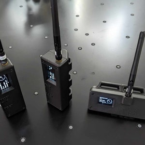 Może przedstawiać: Trzy czarne urządzenia z antenami i małymi ekranami wyświetlającymi tekst i ikonę zegara. Urządzenia znajdują się na czarnej powierzchni z białymi kropkami.