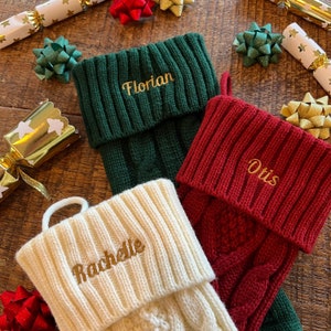 Könnte beinhalten: Drei personalisierte Weihnachtsstrümpfe in Grün, Rot und Creme, jeweils mit einem Namen in Goldfaden bestickt: "Florian", "Otis" und "Rachelle". Weihnachtsknaller und dekorative Schleifen umgeben die Strümpfe.