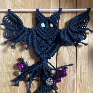 May include: A macrame owl wall hanging in dark blue. The owl has outstretched wings and is adorned with small white beads for eyes. The bottom features purple and silver bells, a pentagram charm, and other decorative elements.