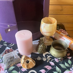 May include: A purple box with a pink candle, a small brass bell, a brown stone, a small bowl, a chalice, a bag of dried herbs, and a stick of incense. The items are arranged on a black and white floral tablecloth.