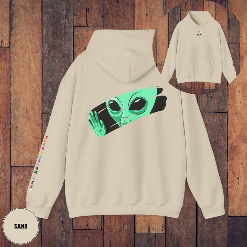Peut inclure: Un sweat-shirt couleur sable avec un visage d'extraterrestre vert qui sort d'un coup de pinceau noir. L'extraterrestre a de grands yeux noirs et porte un gant blanc. Le sweat-shirt a du texte arc-en-ciel sur la manche qui dit "YOUR CUSTOM TEXT".