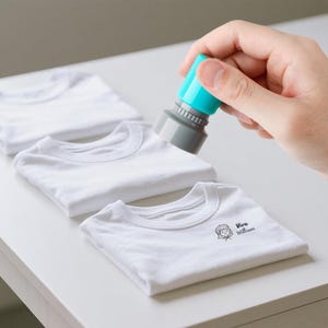 May include: A hand holding a teal and gray stamp over a stack of folded white t-shirts. The bottom shirt has a small graphic and the text "Viva Williams" printed on it.
