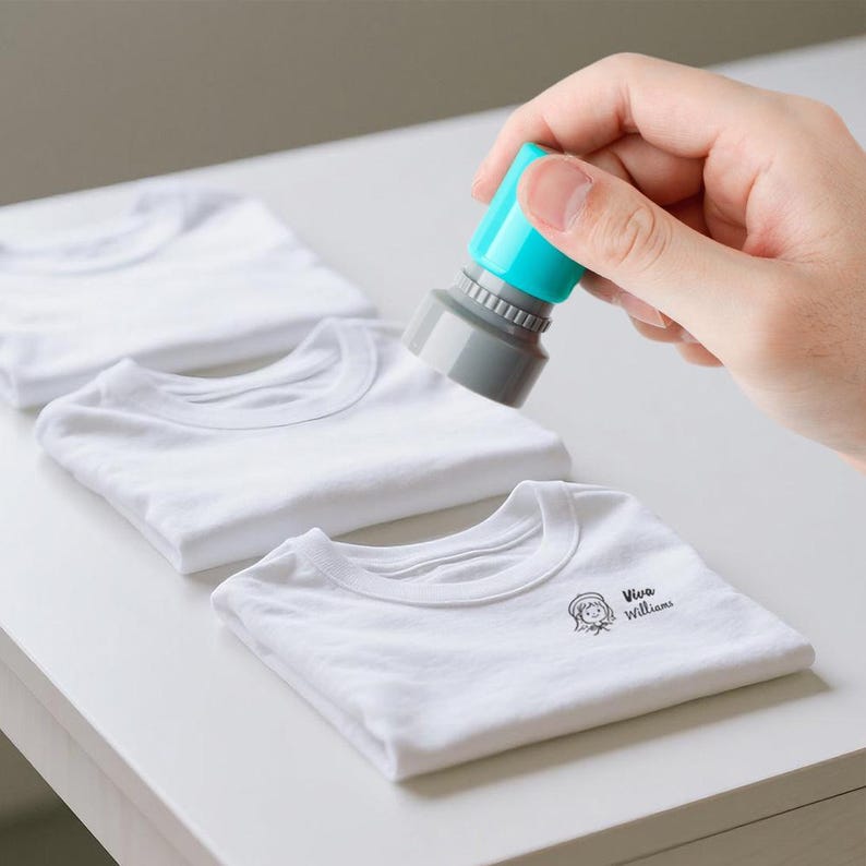 May include: A hand holding a teal and gray stamp over a stack of folded white t-shirts. The bottom shirt has a small graphic and the text "Viva Williams" printed on it.
