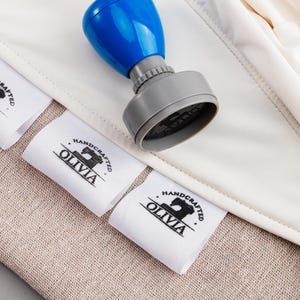 May include: A blue and gray stamp with the text "OLIVIA HANDCRAFTED" printed on white fabric labels. The labels are placed on a beige fabric surface.