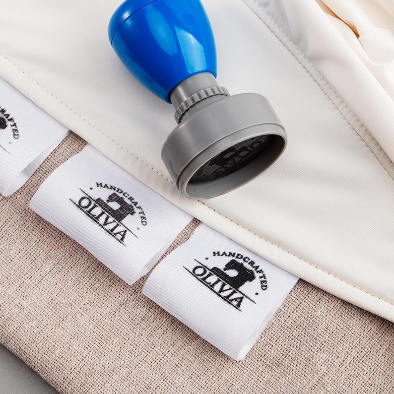May include: A blue and gray stamp with the text "OLIVIA HANDCRAFTED" printed on white fabric labels. The labels are placed on a beige fabric surface.
