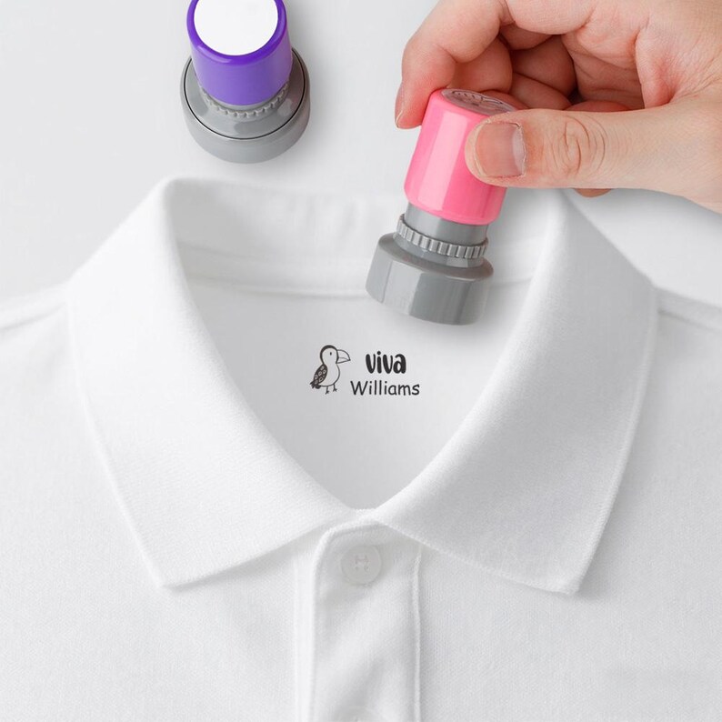 May include: A white polo shirt with a small bird graphic and the text "viva Williams" printed on the chest. Two stamps, one purple and one pink, are positioned above the shirt, suggesting a stamping process.