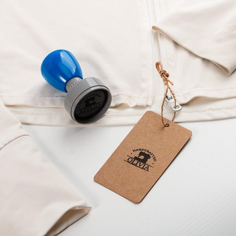 May include: A blue and gray stamp with the text "ALIVIO" and a brown tag with the text "HANDCRAFTED OLIVIA" are displayed on a white garment. The stamp has a blue handle and a gray base. The tag is attached with twine.