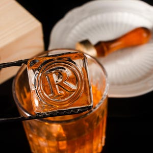 May include: A clear ice cube with the letter "R" embossed inside, held by black tongs over a glass of amber liquid. A wooden box and a white plate with a wooden muddler are in the background. The ice cube is square-shaped.