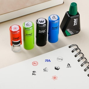May include: A selection of self-inking stamps in red, green, black, and blue, alongside a green-capped ink bottle. The stamps feature various designs. The ink bottle is black. A notepad shows stamped logos. The stamps are for office or craft use.