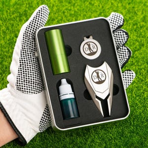 May include: A silver metal golf tool set in a black and silver tin. The set includes a golf ball marker, a divot tool, and a bottle of green liquid. The tin has the letters "D.R." on it.