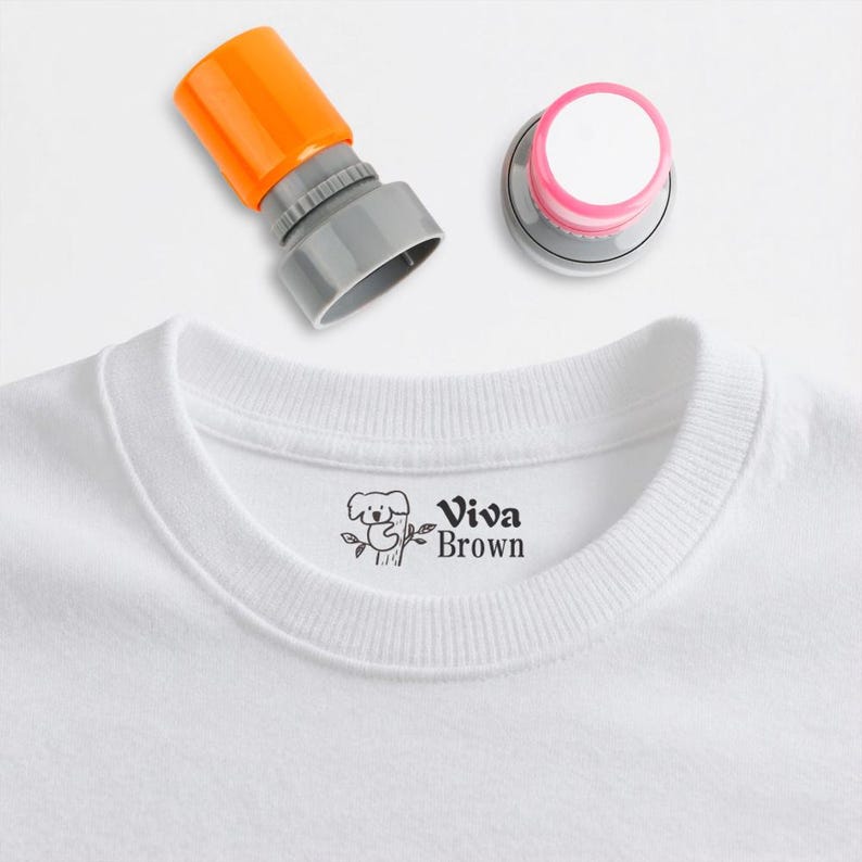 May include: A white t-shirt with a crew neck, featuring a small black graphic of a koala bear and the text "Viva Brown." Two stamps are visible: one with an orange top and the other with a pink top.