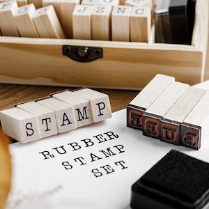 Peut inclure: Un ensemble de tampons en caoutchouc en bois avec des blocs de lettres individuels épelant "STAMP" et "RUBBER STAMP SET". L'ensemble comprend une boîte de rangement en bois et un tampon encreur noir. Les lettres sont dans une police serif classique.