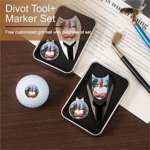 Könnte beinhalten: Ein Golf-Divot-Werkzeug- und -Markierer-Set in einer silbernen Dose mit einem personalisierten Golfball. Das Set enthält ein schwarzes Divot-Werkzeug mit einem silbernen Markierer, ein silbernes Divot-Werkzeug mit einem schwarzen Markierer und einen weißen Golfball mit einem Foto einer Person darauf. Der Text "Divot Tool+ Marker Set" ist oben im Bild gedruckt. Der Text "Free customized golf ball with purchase of set" ist unten im Bild gedruckt.
