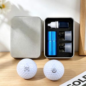 May include: A silver metal box with an open compartment containing golf accessories. Includes a blue and silver container, a small bottle, and two gray pieces. Two white golf balls with logos are in front of the box.