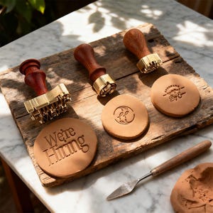 Könnte beinhalten: Eine Sammlung von Stempeln mit Holzgriffen und Messingstempelköpfen, zusammen mit gestempelten Lederkreisen. Ein Kreis trägt die Aufschrift "We're Hiring". Ein kleiner Metallspatel und Ton sind ebenfalls vorhanden.