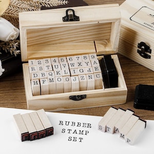 May include: A wooden rubber stamp set with individual letter and number stamps, stored in a wooden box. The set includes an ink pad and the words "RUBBER STAMP SET" printed on a white surface. The box is open, revealing the stamps.