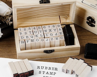 May include: A wooden rubber stamp set with individual letter and number stamps, stored in a wooden box. The set includes an ink pad and the words "RUBBER STAMP SET" printed on a white surface. The box is open, revealing the stamps.