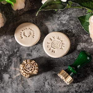 May include: A collection of pottery stamps and clay pieces. Two round clay pieces are stamped with text and a deer antler design and a rose. A brass stamp and a green handled stamp are also present.