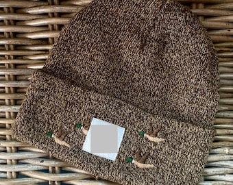 May include: Brown knit beanie with embroidered ducks. The hat features a folded brim and a square patch. The ducks are embroidered in shades of brown, green, and white. The beanie is made of a textured knit material.