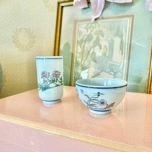May include: Two small white ceramic bowls with floral designs. The bowl on the left is a vase with a narrow opening and the bowl on the right is a wider bowl. Both bowls have blue trim and feature pink and green floral designs.
