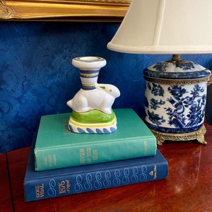 May include: A white ceramic candlestick shaped like a rabbit with green, yellow, and blue accents. The candlestick is sitting on top of a stack of three books. The top book is green and has the title "River of the Sun" on the spine. The middle book is blue and has the title "1876" on the spine. The bottom book is blue and has the title "Gore Vidal" on the spine.